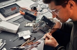 A man wearing glasses and gloves repairs a smartphone at a cluttered desk. Tools, a magnifying glass, and electronic components are scattered around.